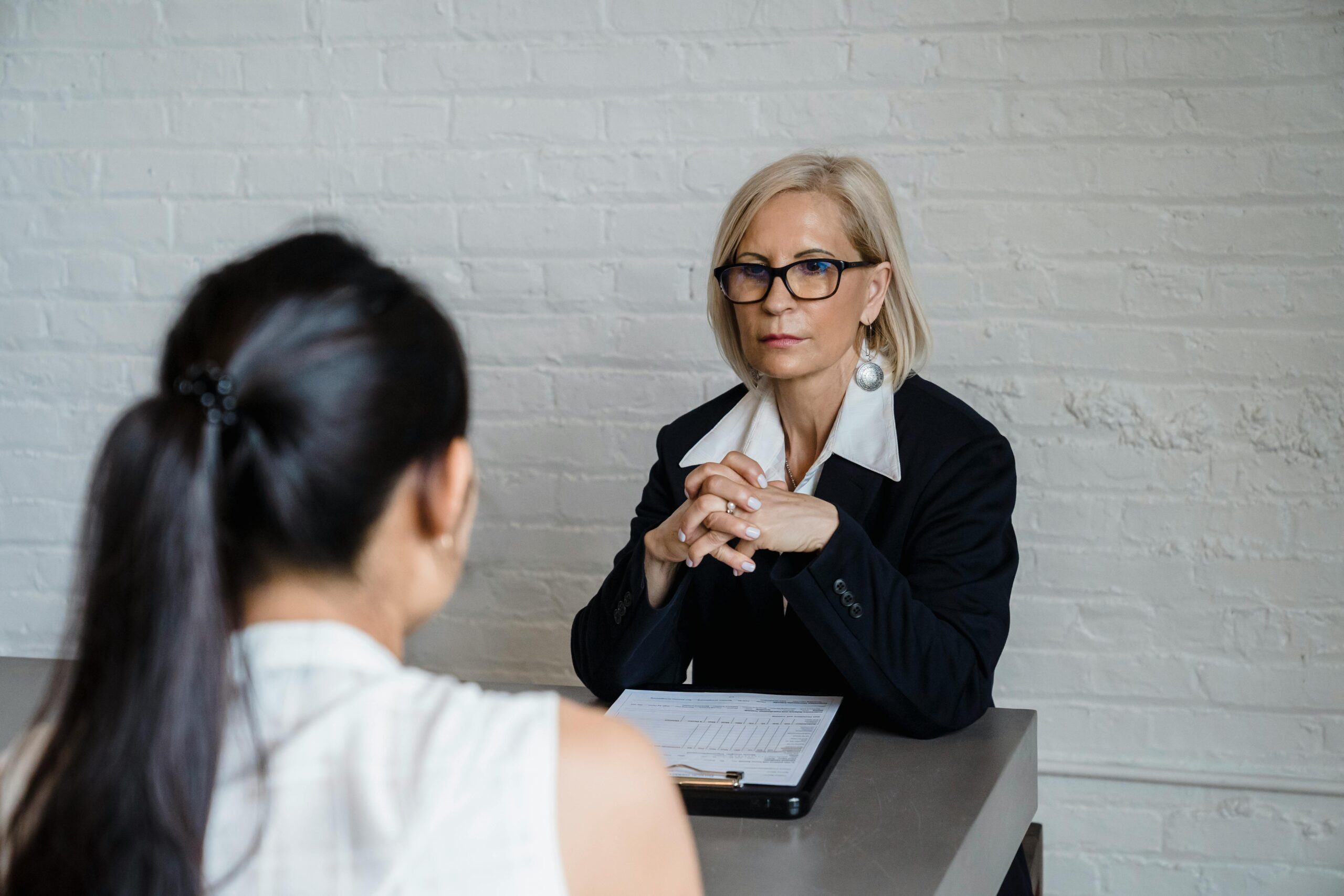 L'entretien structuré pendant un recrutement : Entretien professionnel entre deux femmes dans un bureau, l’une prenant des notes sur un clipboard, l’autre écoutant attentivement, avec un mur en briques blanches en arrière-plan.